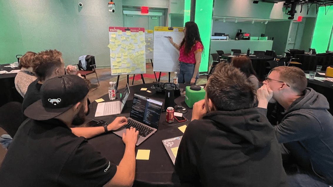 CSS, UI, and DevTools team sit around the table in a conference room. Una stands at a whiteboard which is covered in sticky notes. Other team members sit around the table with refreshments and laptops.