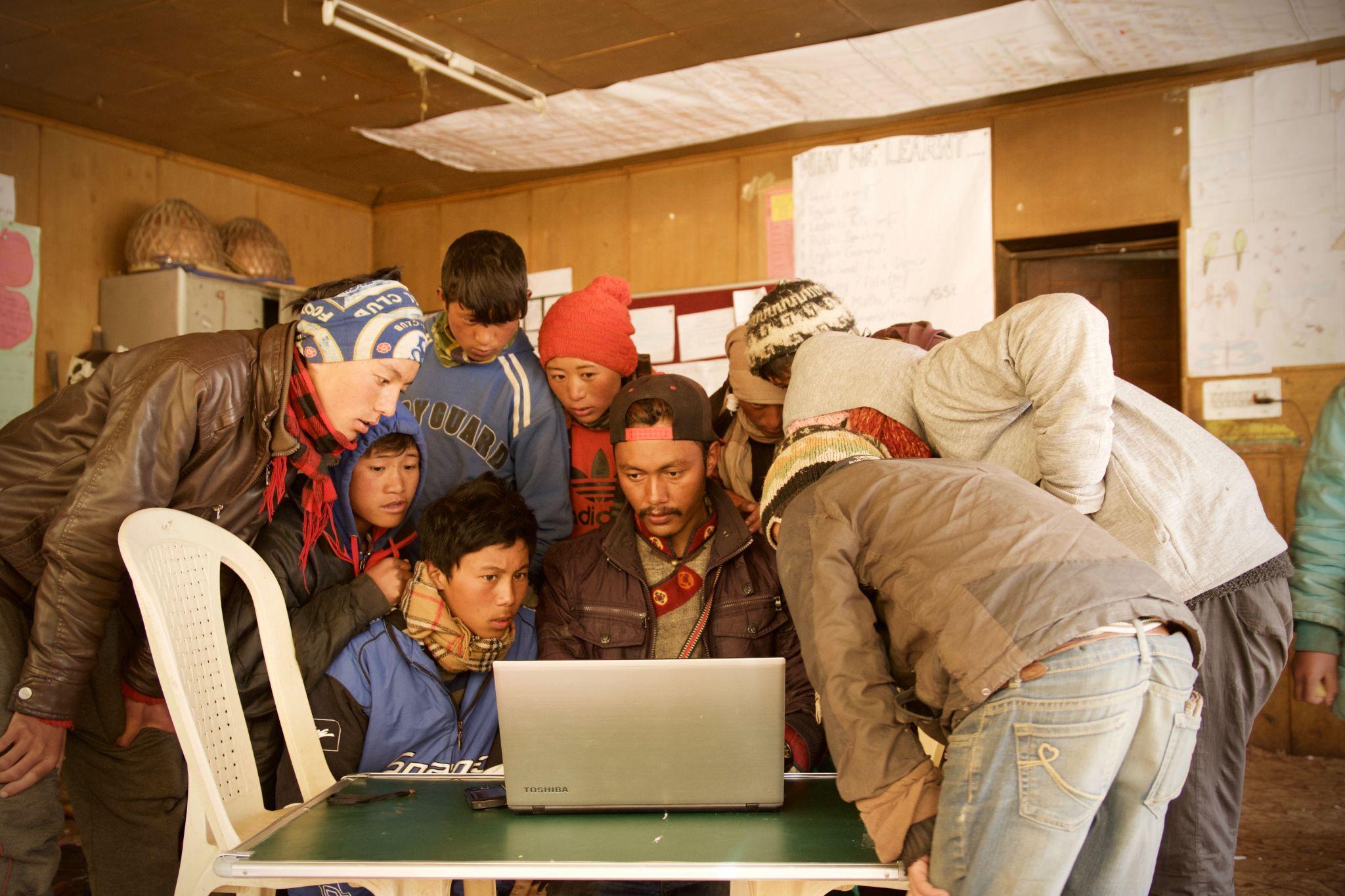 Люди собираются вокруг ноутбука, стоящего на простом столе с пластиковым стулом слева. Фон выглядит как школа в развивающейся стране.
